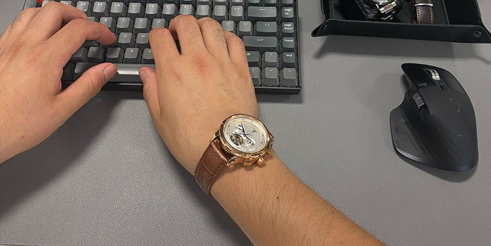 Person typing on a keyboard, wearing a watch with a brown leather strap.
